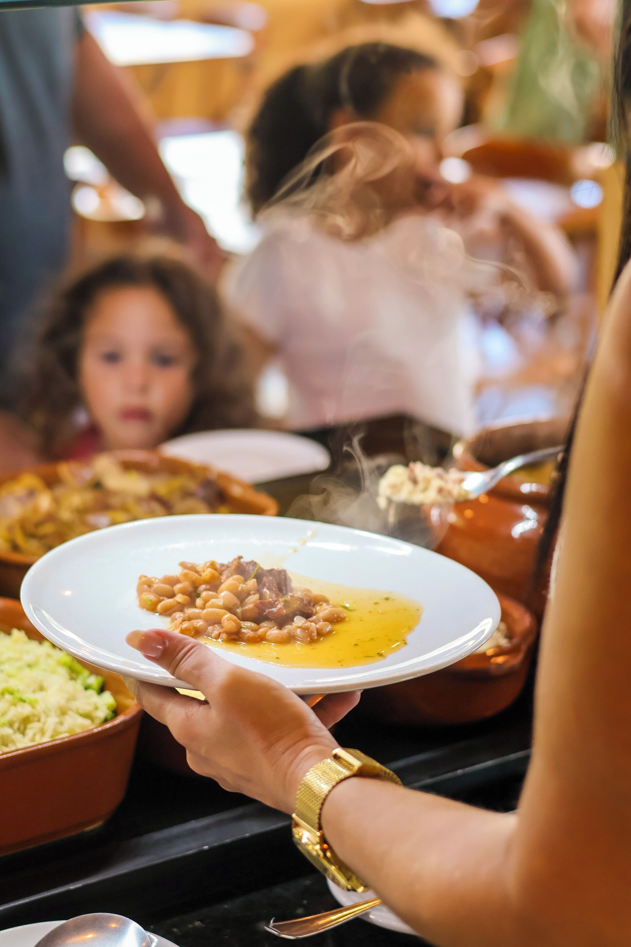 A person holding a plate with beans and meat, with other dishes and people in the background.