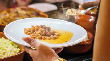 A person holding a plate with beans and meat, with other dishes and people in the background.