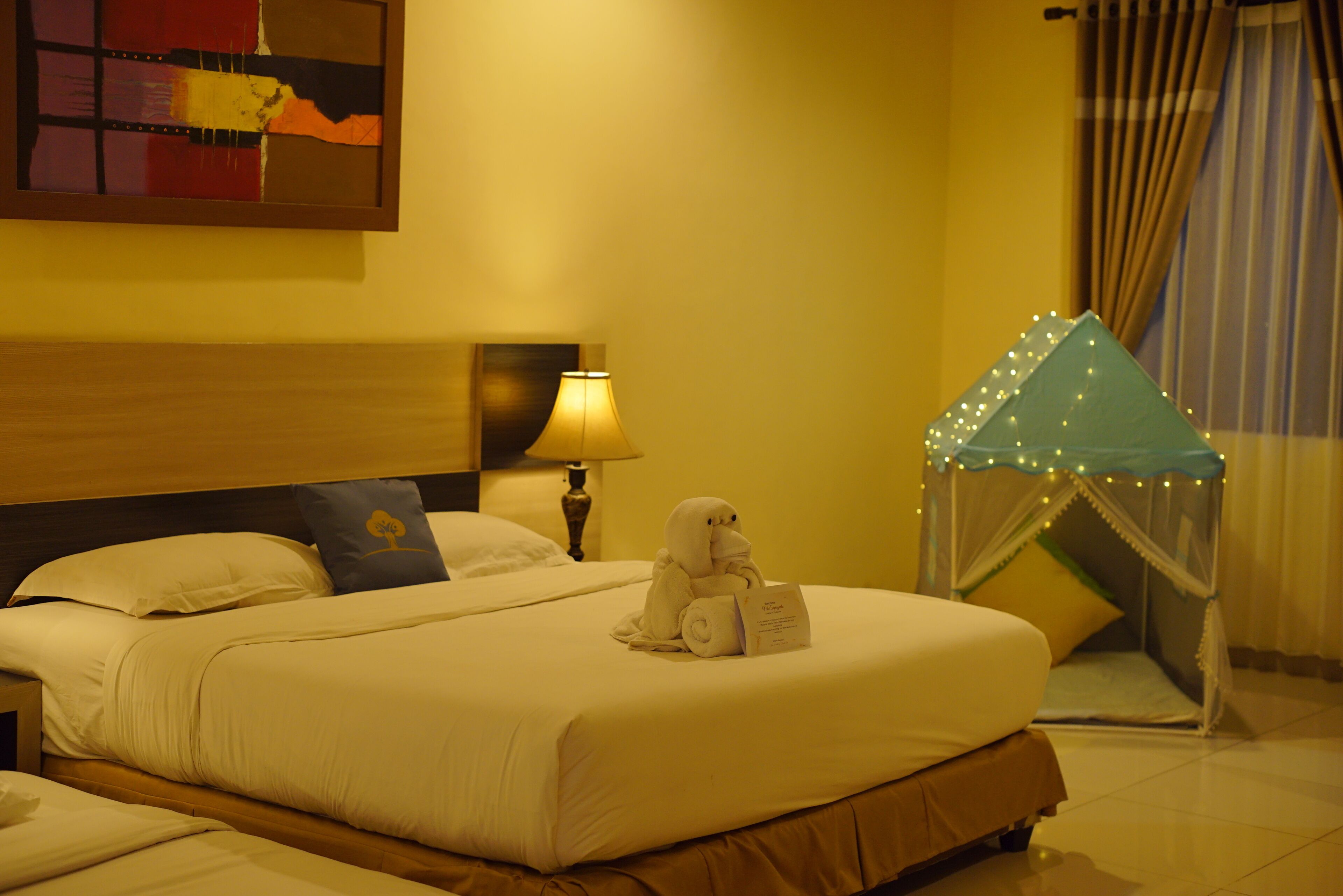 Habitación de hotel con una cama grande, una mesita de noche con lámpara, un elemento decorativo en la pared y una cama con dosel e iluminación.