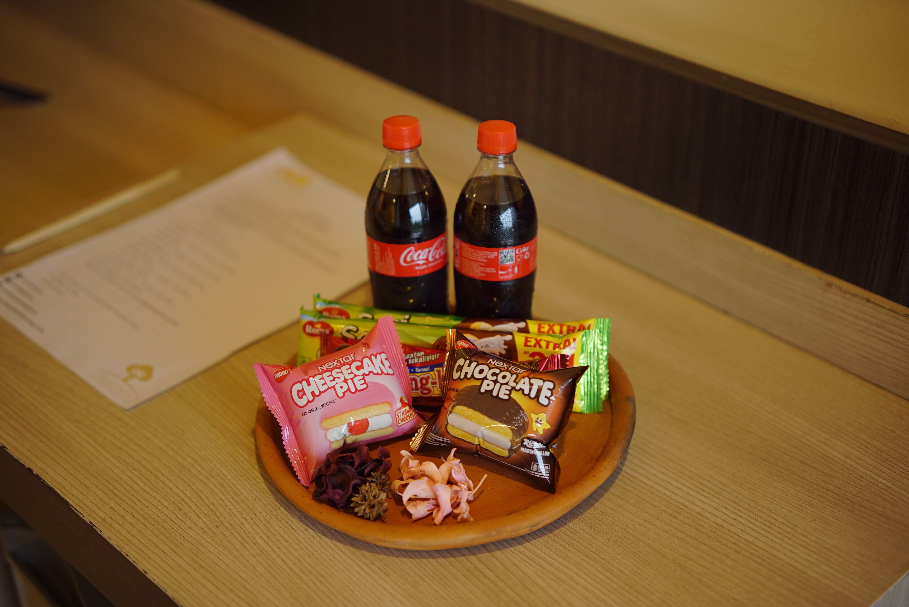 A wooden table with a plate of snacks, two bottles of Coca-Cola, and two packages of chocolate and cheesecake pies.
