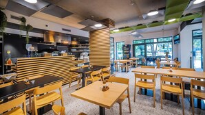 A modern restaurant interior with wooden furniture, a pizza counter, and large windows offering a view of the outdoors.