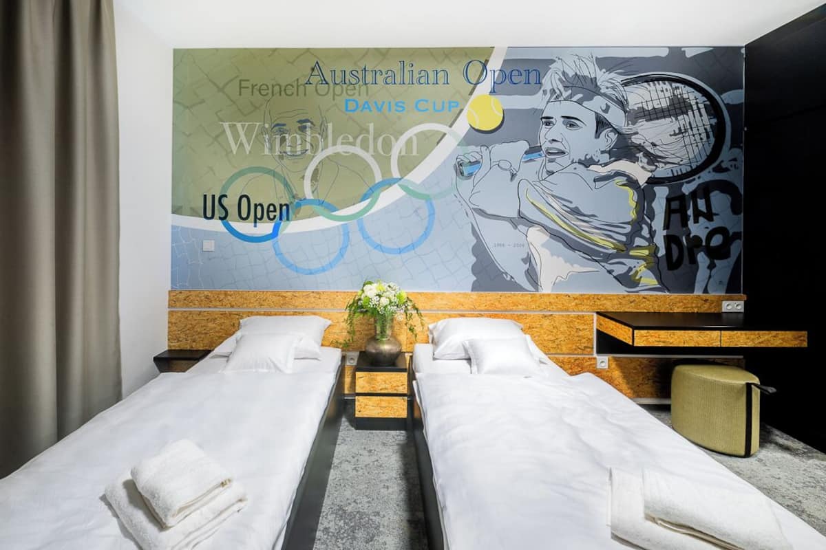 Une chambre d’hôtel avec deux lits, une grande fresque murale représentant des joueurs de tennis et une petite table sur laquelle est posé un vase de fleurs.