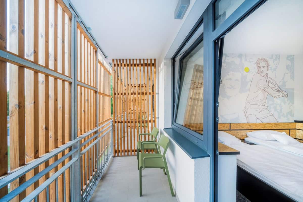 Un couloir étroit, avec une clôture en bois d’un côté et un lit près d’une fenêtre de l’autre.