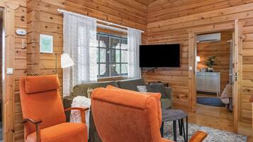 Een gezellig interieur van een blokhut, met houten wanden, een aan de wand bevestigde televisie en een zithoek met twee oranje fauteuils en een bank.