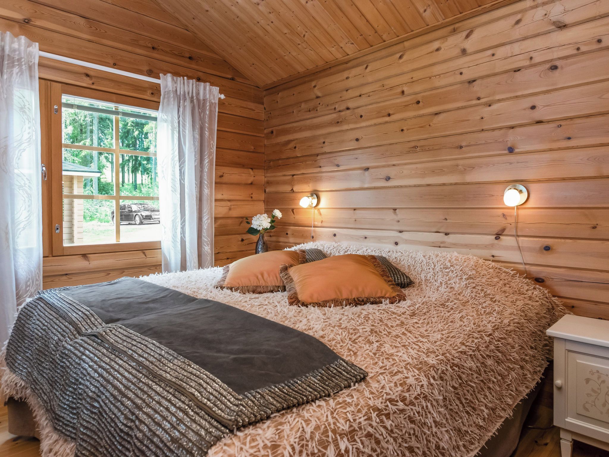 A wooden cabin bedroom with a bed, pillows, a nightstand, and a window with curtains.