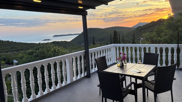 Un balcone con tavolo e sedie, affaccia su un paesaggio costiero con colline e vista sul mare.