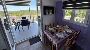 Un'area pranzo con un tavolo apparecchiato per due, una televisione montata a parete e un balcone con vista su dolci colline.