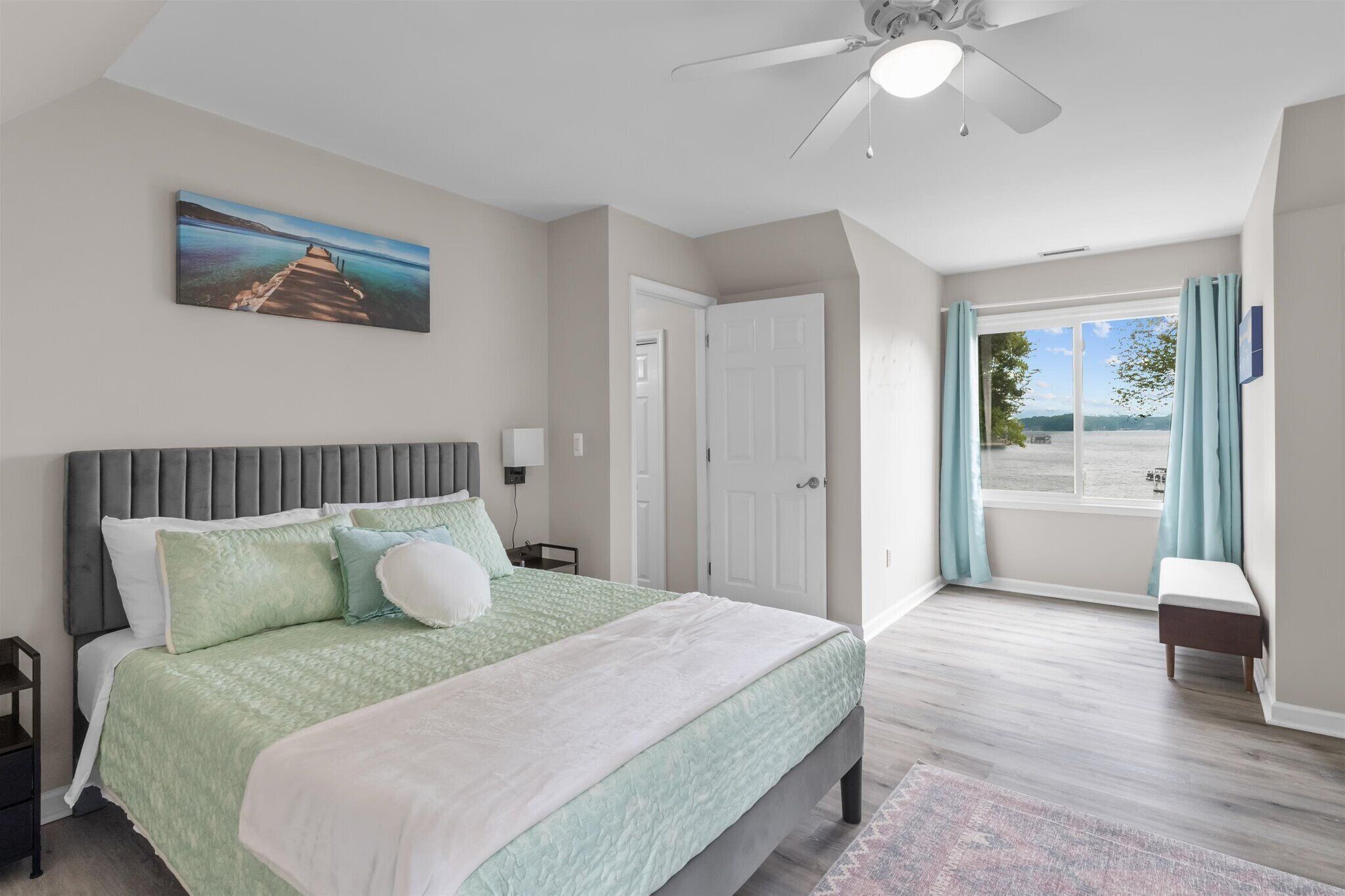 A bedroom with a bed, a ceiling fan, a painting, a door, and a view of the outdoors.