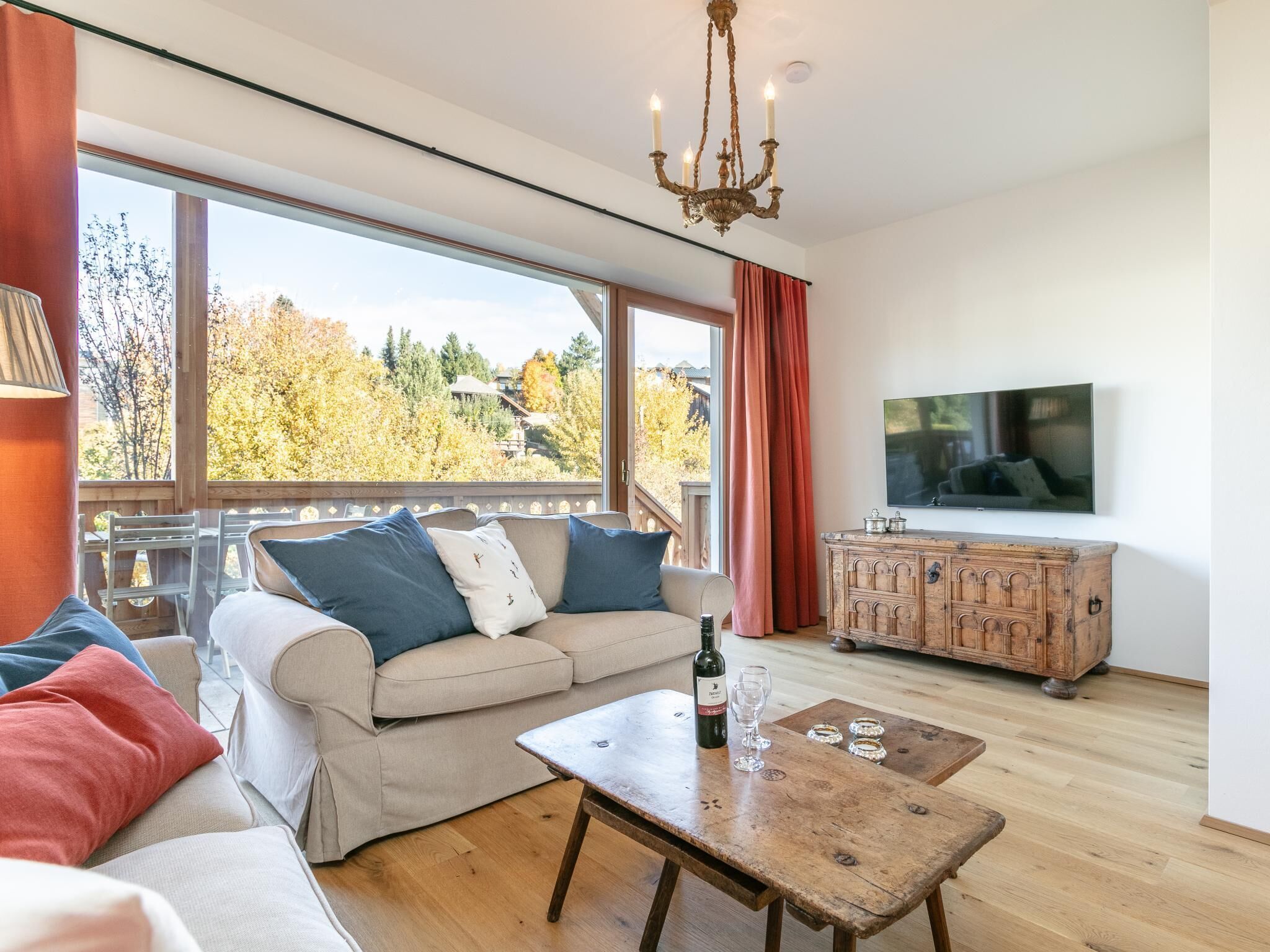 Una sala de estar acogedora con un sofá, una mesa de centro de madera, un televisor y vistas a árboles otoñales a través de amplios ventanales.