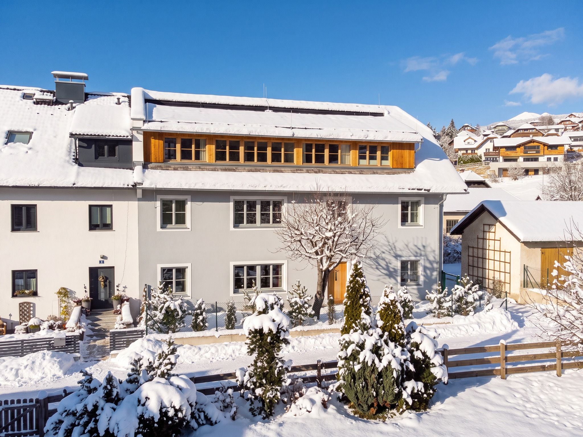 Un edificio de varios pisos con tejado cubierto de nieve y un jardín con árboles nevados.