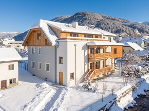 A two-story chalet with a wooden balcony and a snow-covered roof, surrounded by a mountainous landscape.