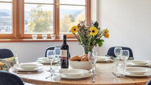Una mesa de comedor dispuesta con platos, vasos, una botella de vino, un jarrón con flores y un cesto de pan.