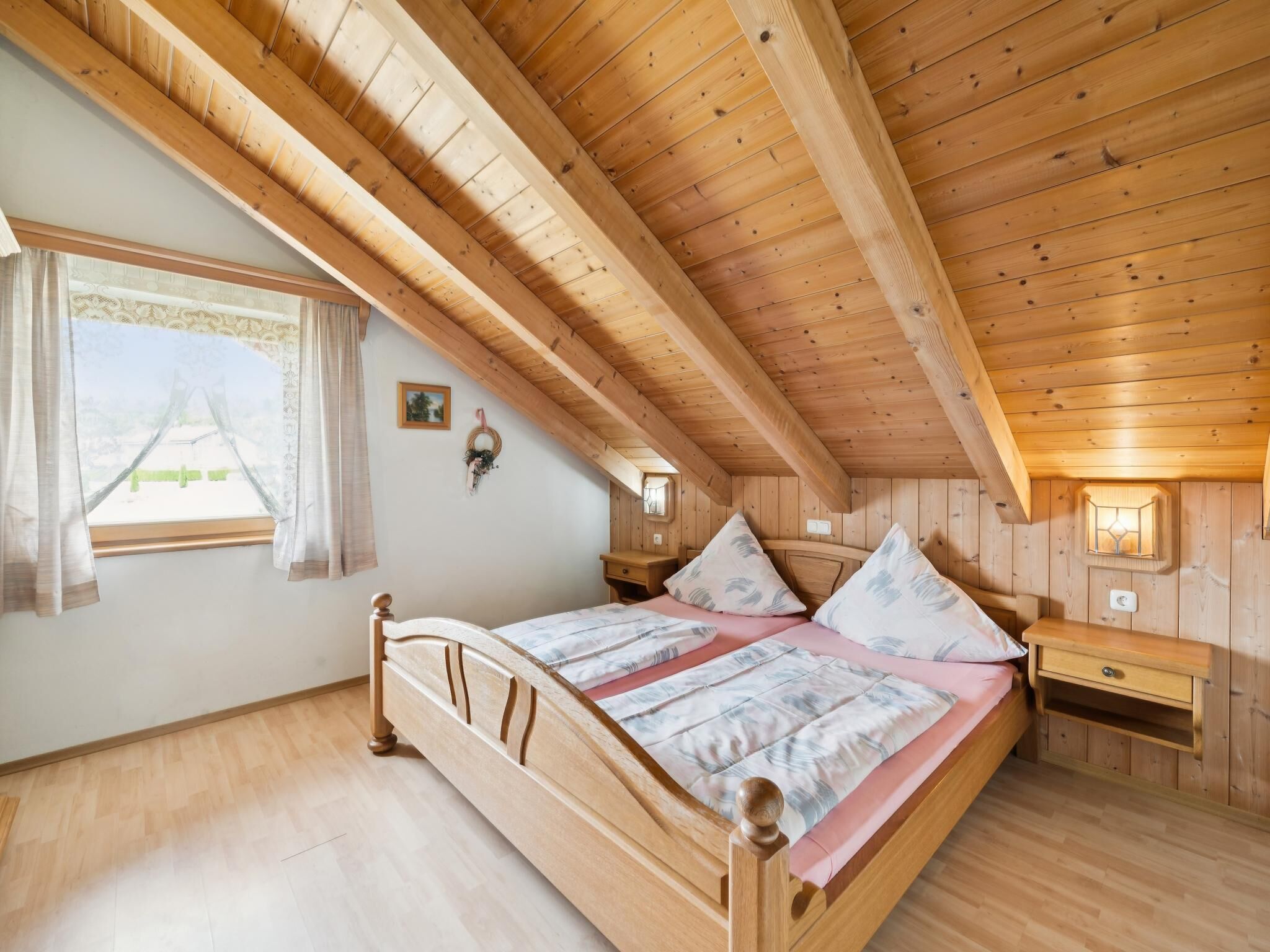 A wooden bedroom with a sloped ceiling, a bed with a patterned bedspread, a nightstand, and a wall-mounted lamp.