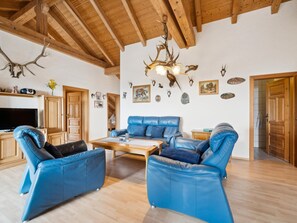 A spacious living room with a high wooden ceiling, a blue leather sofa set, a wooden coffee table, and various wall-mounted animal horns and antlers.