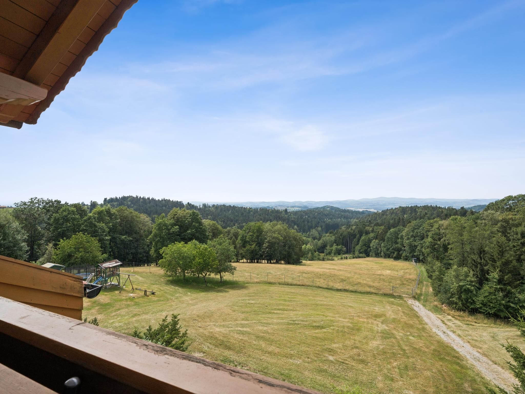 Vista desde un balcón sobre un campo cubierto de hierba, con árboles y colinas al fondo.