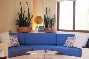 A blue sofa with white and blue patterned cushions, a round white coffee table, and two large potted plants with long green leaves.