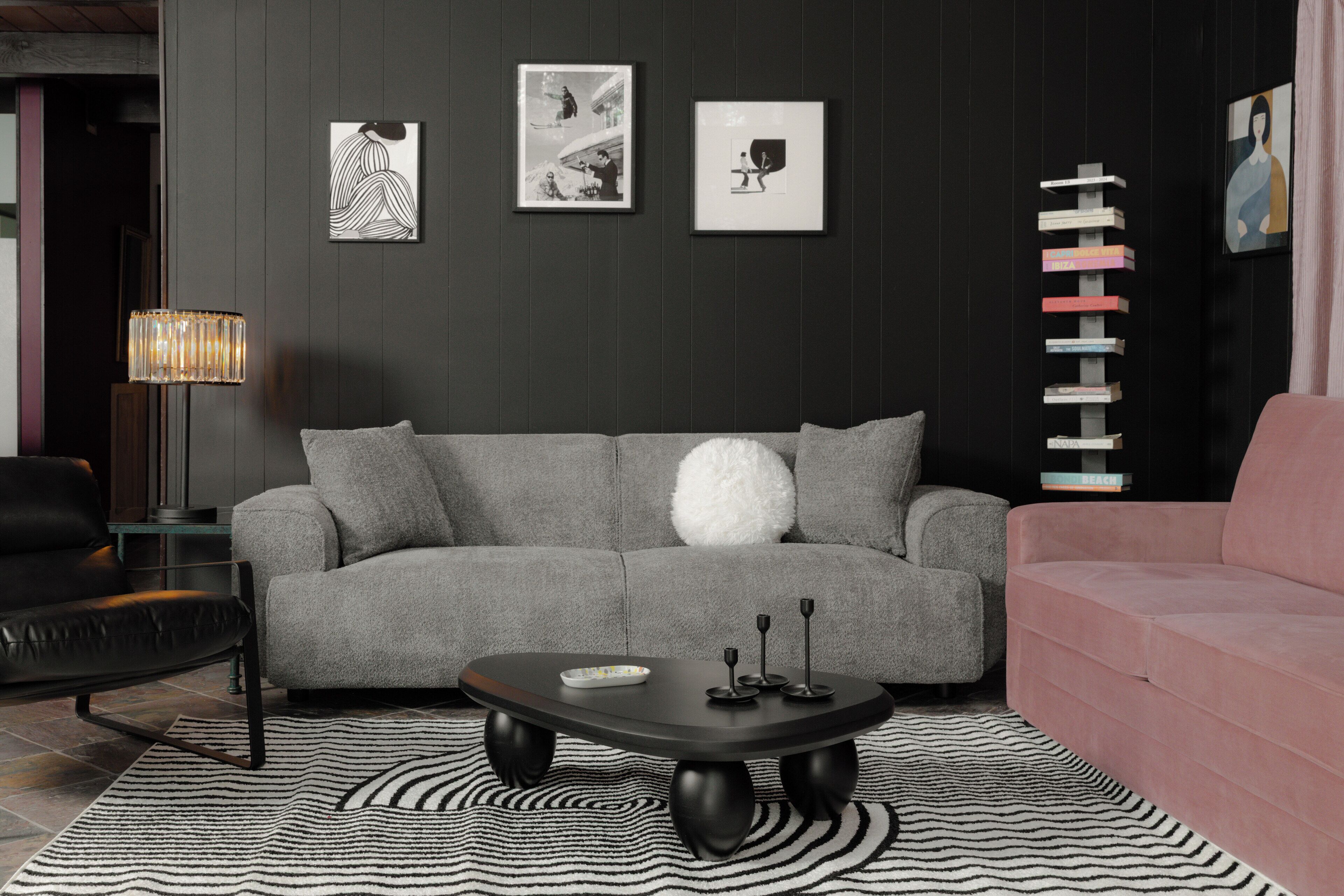 Een moderne woonkamer met een grijze bank, een zwarte salontafel en een vloerkleed met zebraprint. Er hangen ingelijste kunstwerken aan de muur en er staat een hoge, kleurrijke boekenkast.