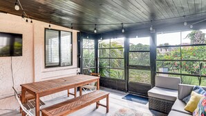Quarto com mesa de madeira e bancos, um sofá, tapete estampado e janelas amplas com vista para o verde.