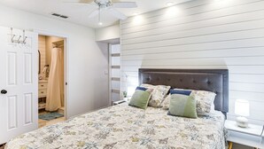 Quarto com cama, mesa de cabeceira, ventilador de teto e banheiro visível por uma porta aberta.