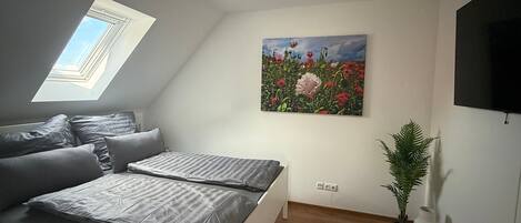 A bedroom with a skylight, a bed with grey bedding, a painting of flowers, a TV, and a potted plant.