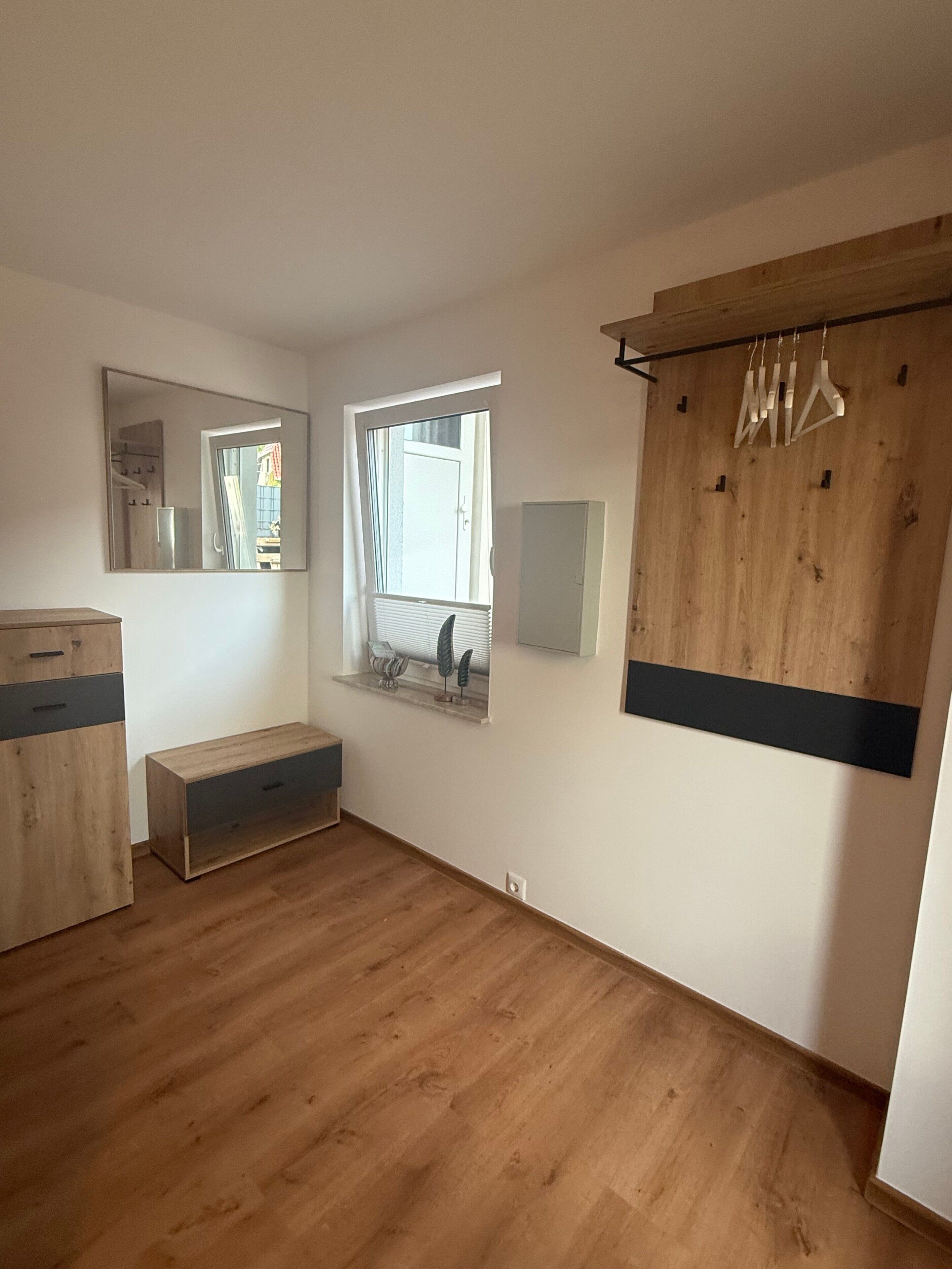 Una habitación con piso de madera, una ventana con persianas, un espejo, un armario de madera y un perchero con colgadores.