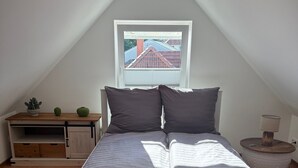 A bedroom with a sloped ceiling, a bed with grey bedding, a small bedside table with a lamp, and a window with a view of buildings. - Spacious Apartment with Lake View, Shared Terrace and Wi-Fi (Feldberger Seenlandschaft)