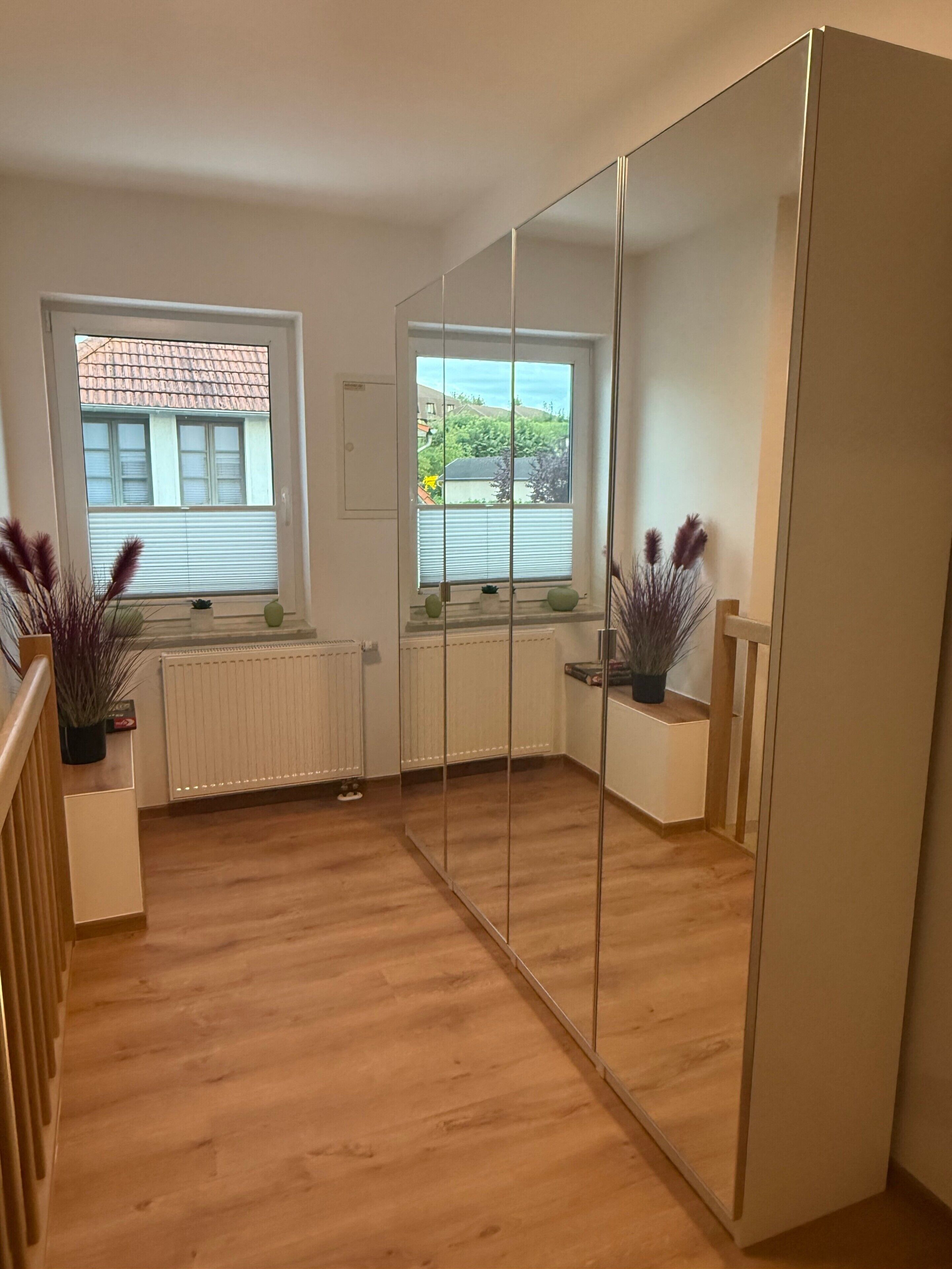 A room with a large mirror, wooden flooring, a radiator, and a window with blinds.