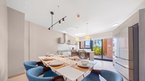 Un espace de repas moderne avec une longue table dressée pour six personnes, des chaises rembourrées bleues et, au fond, une cuisine donnant sur un paysage verdoyant.