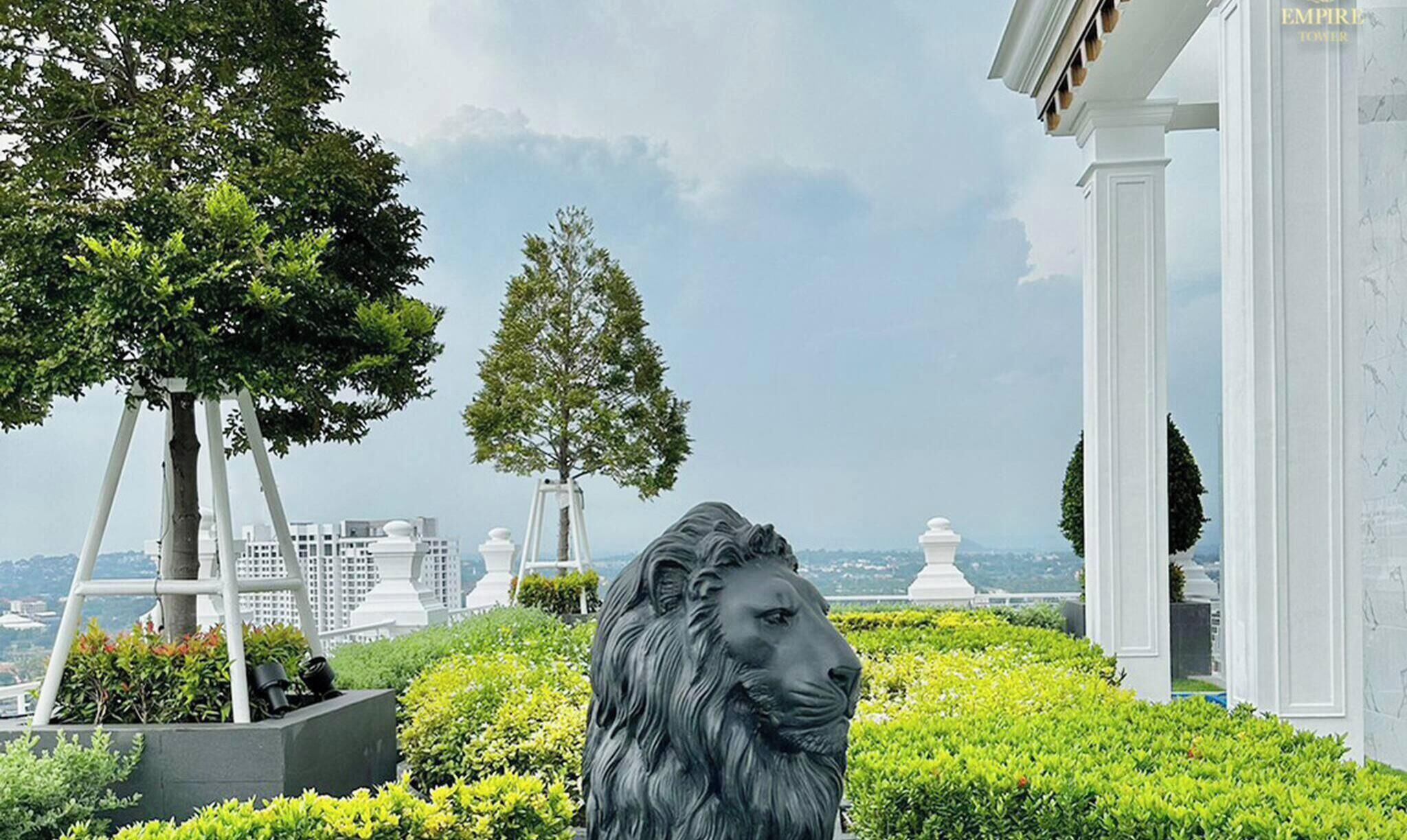 A statue of a lion in a landscaped area with trees and a view of buildings in the background.