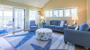 A living room with a sofa, armchairs, a coffee table, and a patterned ottoman. There are two windows and a door leading to a balcony.