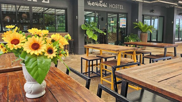 El interior de un hostal muestra mesas y bancos de madera, un ventanal grande y un cartel que dice "HOSTEL".