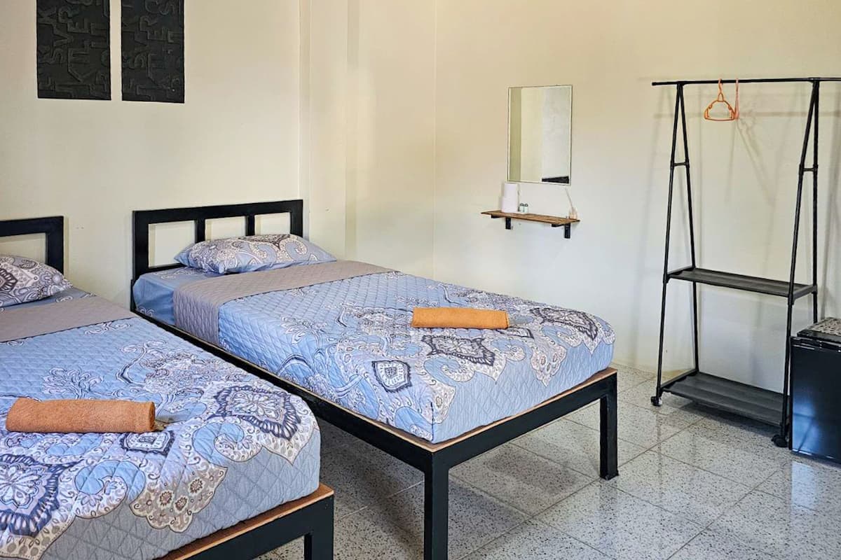 A room with two beds, each with patterned bedding and a rolled towel. There is a mirror on the wall and a shelf with items.