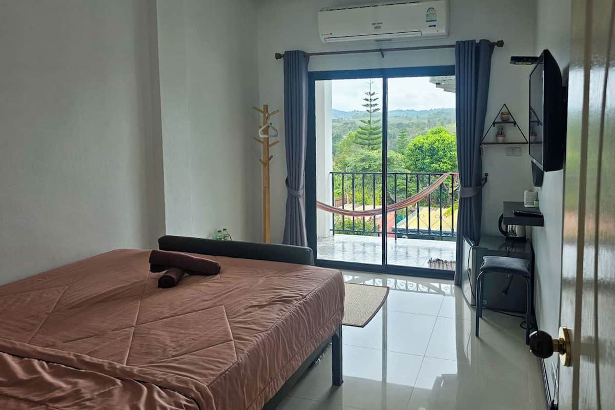 A hotel room with a bed, a TV, a balcony with a hammock, and a view of greenery.
