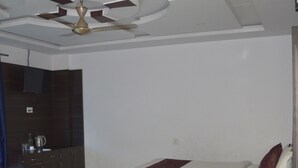 Een moderne hotelkamer met een bed, een blauwe bank, een houten salontafel, een plafondventilator en een kitchenette met kasten en een televisie.
