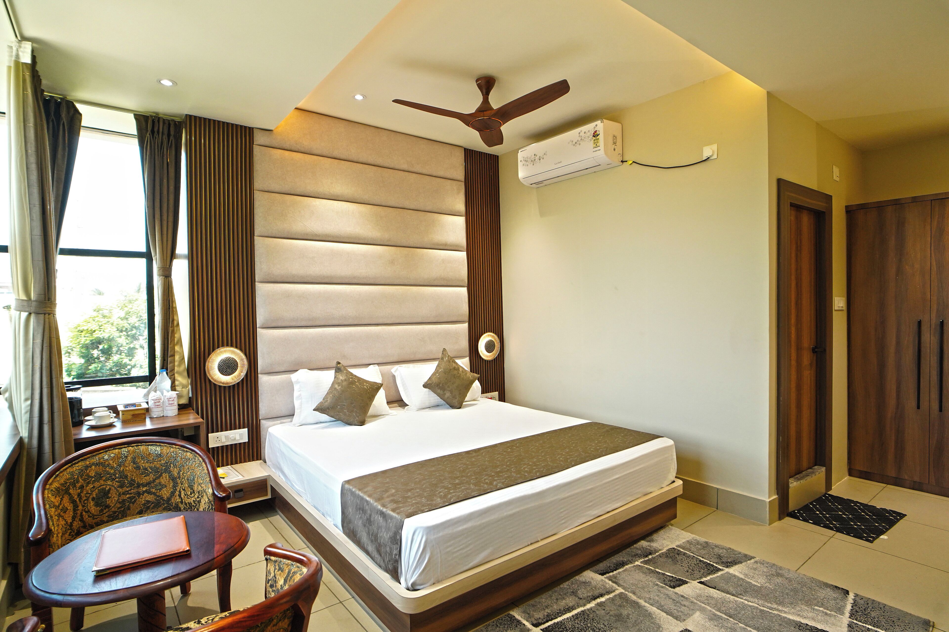 A hotel room with a large bed, a desk with a chair, a ceiling fan, and an air conditioning unit.