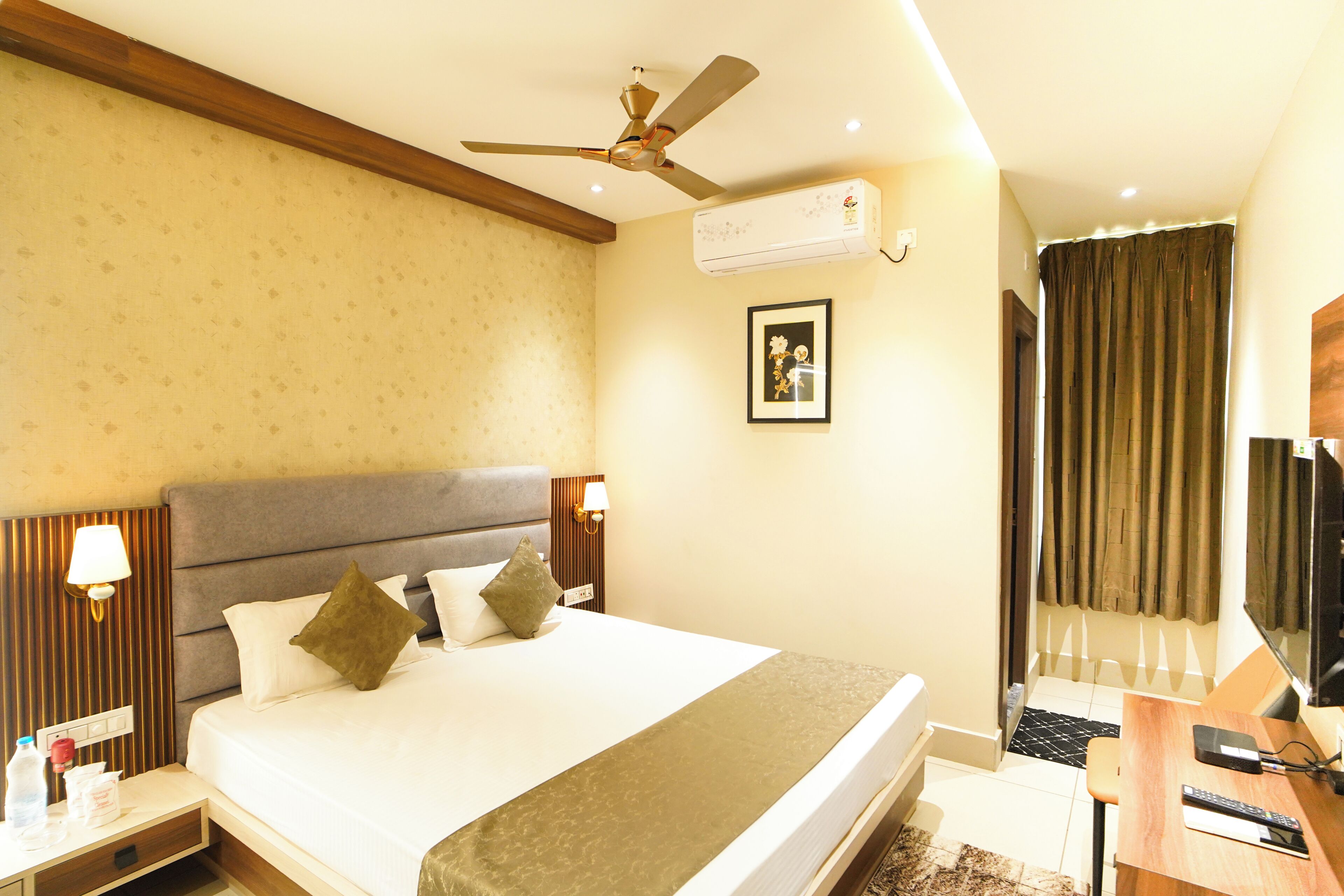 Quarto de hotel com cama, mesa de cabeceira, luminária, ar condicionado, ventoinha no teto e televisão.