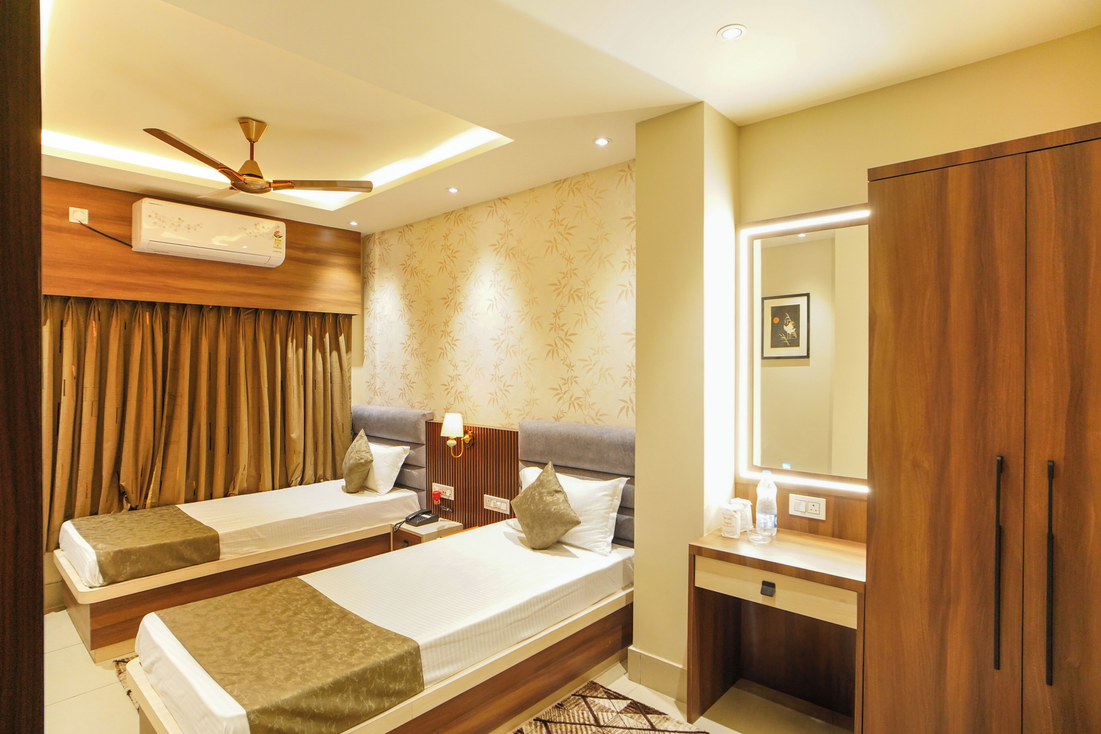 A hotel room with two beds, a ceiling fan, an air conditioning unit, a bedside table with a lamp, a mirror, and a wardrobe.