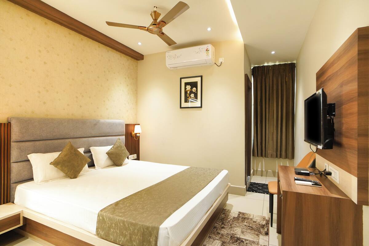 A hotel room with a bed, a desk, a television, a ceiling fan, an air conditioning unit, and a framed picture on the wall.