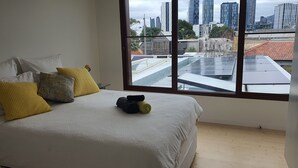A bedroom with a large window, a bed with a white duvet, and a view of a cityscape with tall buildings and solar panels on the roof.