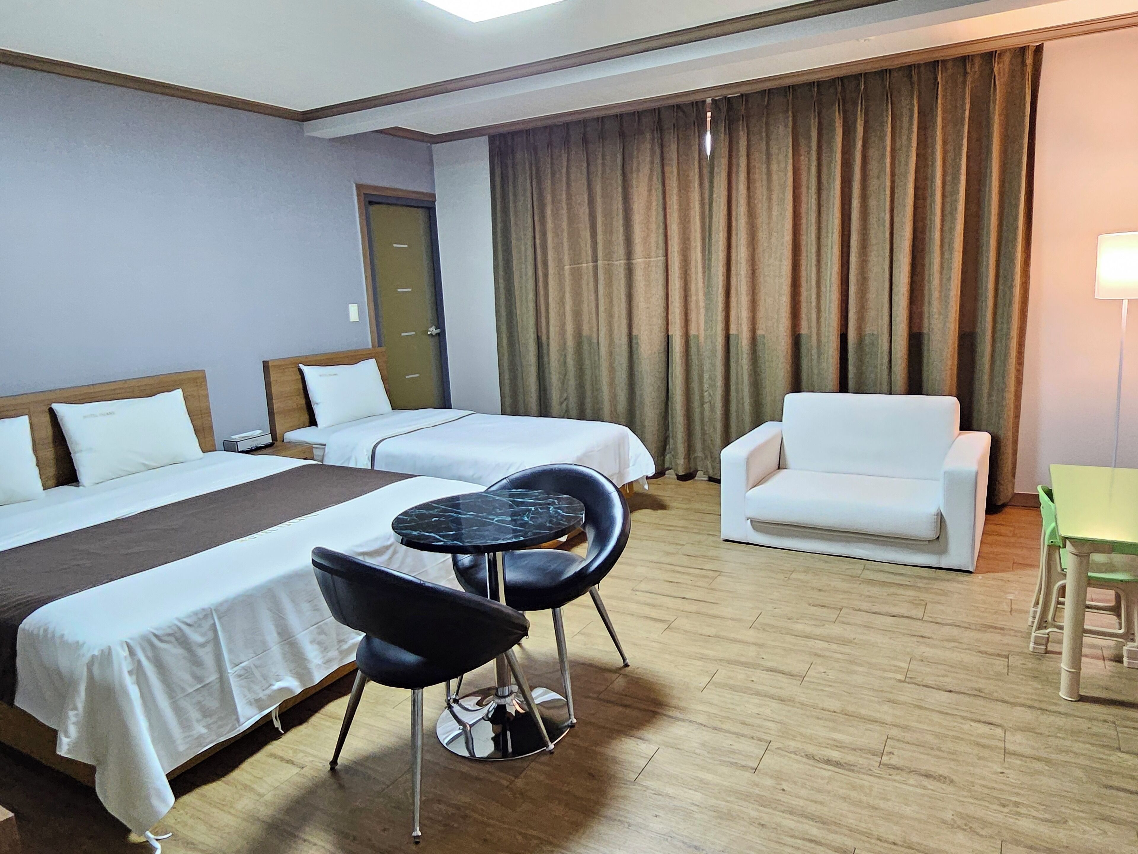 Una habitación de hotel con dos camas, una mesita redonda pequeña, una silla negra, un sillón blanco y una mesa verde.