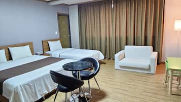 Una habitación de hotel con dos camas, una mesita redonda pequeña, una silla negra, un sillón blanco y una mesa verde.
