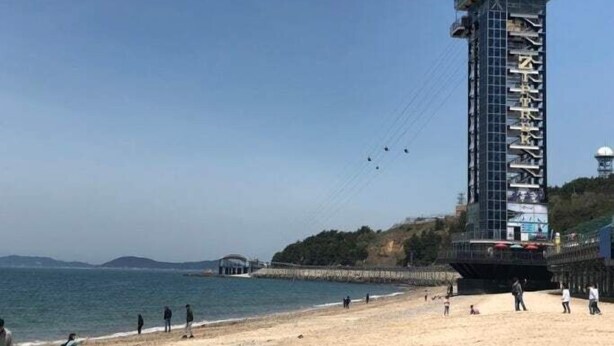 Una torre alta y moderna con una plataforma panorámica, ubicada en una playa con vistas despejadas al mar y colinas distantes.