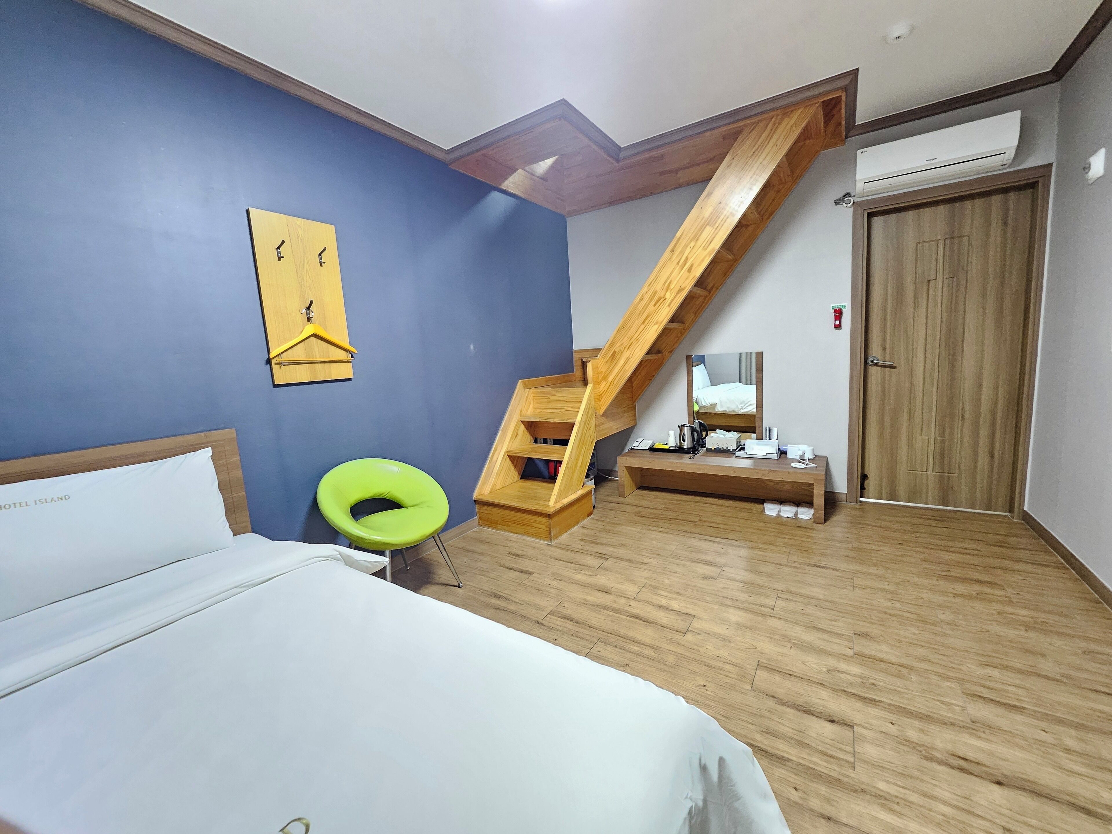 Ein Schlafzimmer mit einem Bett, einem grünen Sessel, einem Holztisch, einem Spiegel, einer Treppe und einer Tür.
