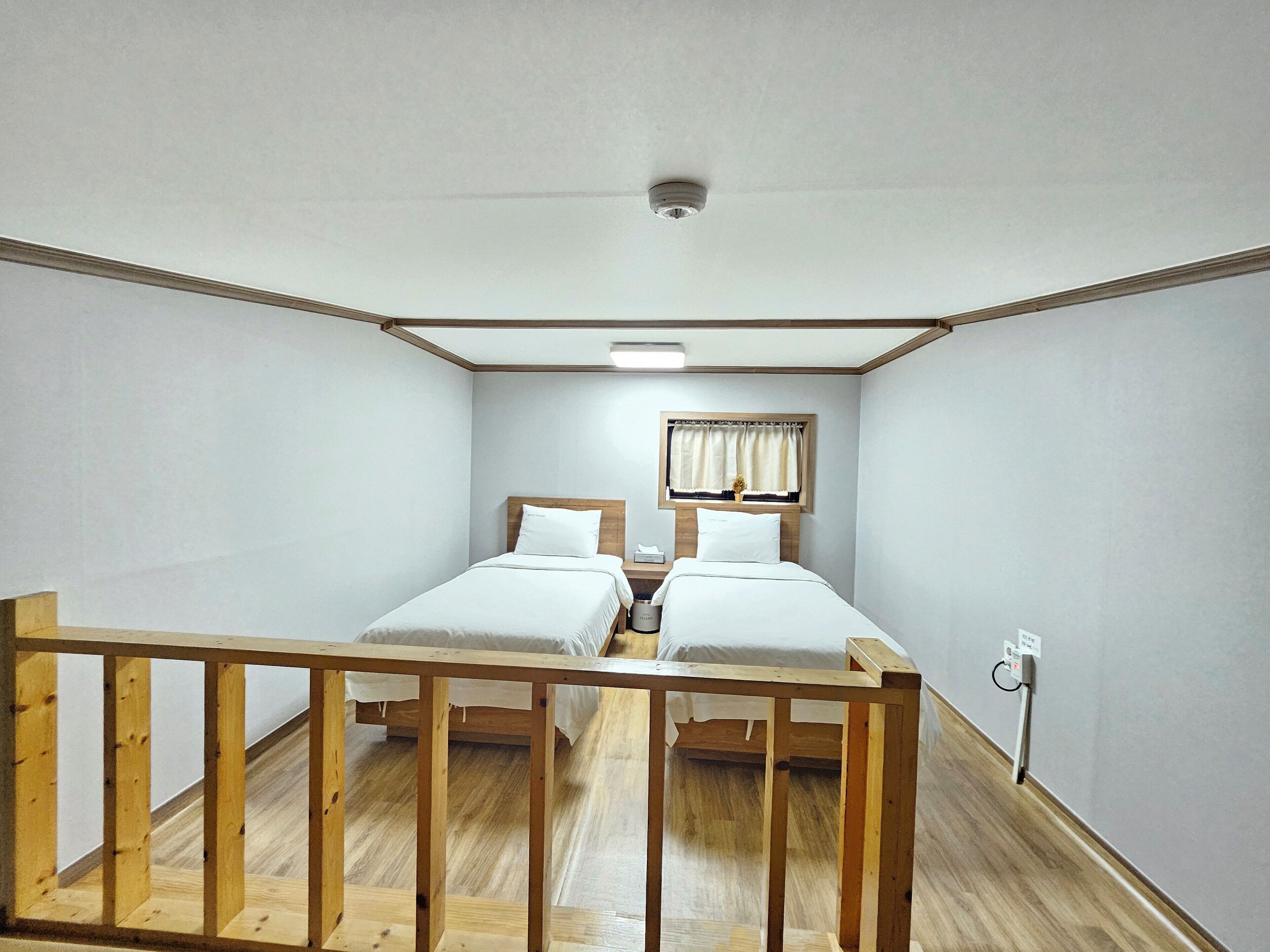 A small, well-lit room with two beds, a window with curtains, and a wooden railing.