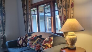 Un salón con un sofá azul, cojines estampados, una mesita de madera con una lámpara y cortinas florales.