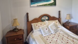 Un dormitorio con cabecera de madera, un cuadro de una playa, dos mesitas de noche con lámparas y ropa de cama estampada con motivos florales.