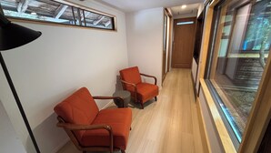 A narrow hallway with two orange armchairs, a round side table, and a large window with a view of a wooden structure.