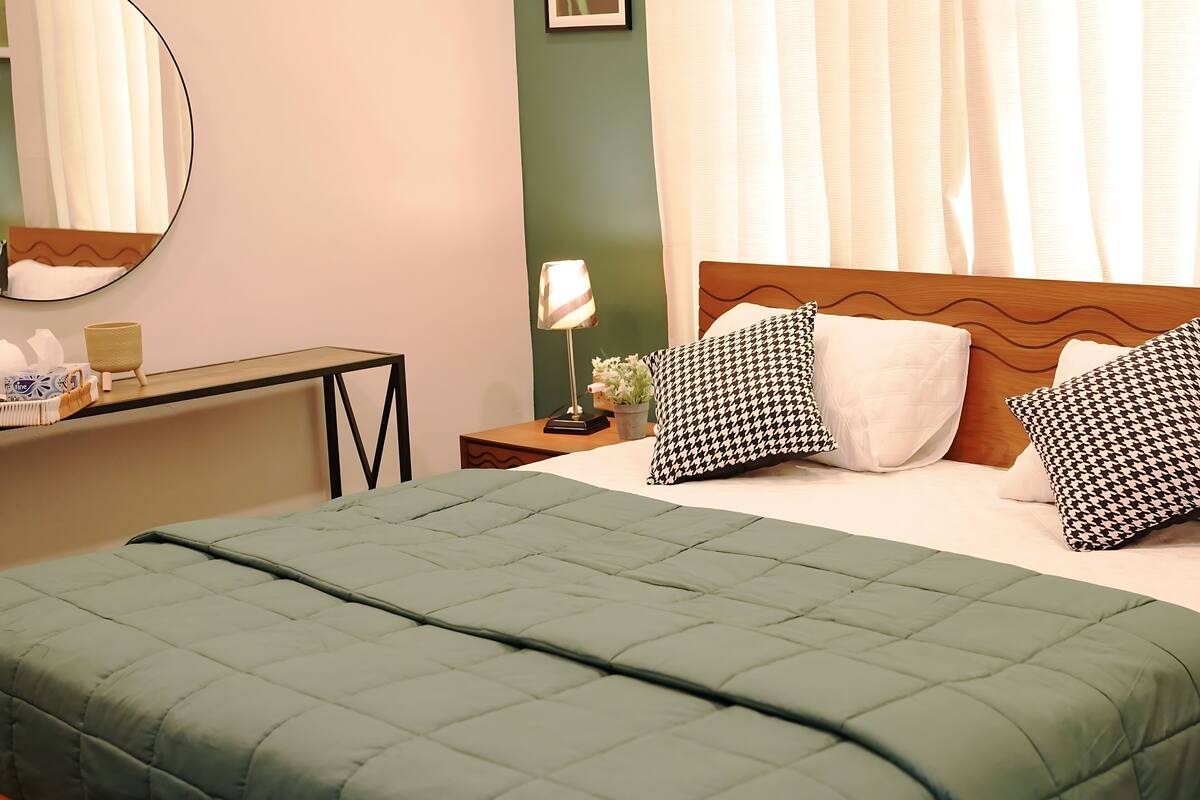 Un lit bien fait, avec une couette verte, une lampe de chevet et des coussins à motifs.