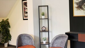 A modern interior with two grey upholstered chairs with orange cushions, a small round table, a tall black shelving unit with potted plants, and a framed picture on the wall.