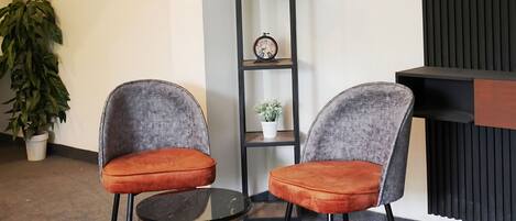 A modern interior with two grey upholstered chairs with orange cushions, a small round table, a tall black shelving unit with potted plants, and a framed picture on the wall.